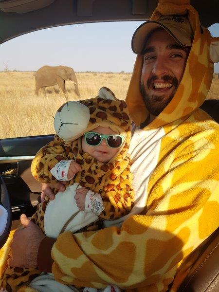 Dad in giraffe onesie holding baby in leopard suit with elephant in background